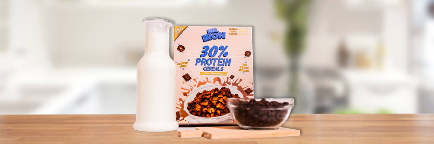 Cutie de cereale proteice Mr. Iron cu 30% proteine si ciocolata fina, alaturi de un bol cu cereale si o sticla de lapte alb, pe un blat de lemn intr-o bucatarie luminoasa.
